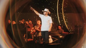 El cantante Edén Muñoz sonríe y señala al público durante un concierto, acompañado por su banda de músicos bajo luces cálidas y un efecto de halo.