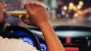 Se ve a un hombre tomando un líquido de una botella y él viste una playera blanca; está dentro de un auto.