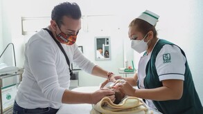 Una enfermera del IMSS y un padre de familia, ambos con mascarillas, atienden a un recién nacido sobre una mesa de auscultación en un consultorio.