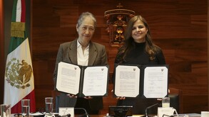 Dos mujeres muestran documentos oficiales firmados frente al escudo de la Universidad de Guadalajara, con una bandera de México al costado.