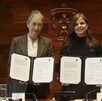 Dos mujeres muestran documentos oficiales firmados frente al escudo de la Universidad de Guadalajara, con una bandera de México al costado.