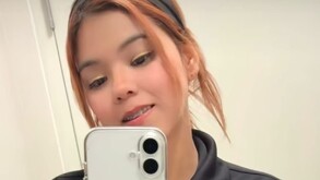 Se observa a una mujer con una diadema en el cabello, viste una chamarra negra y trae un celular blanco en la mano.