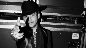 El cantante Christian Nodal, en una foto a blanco y negro, viste sombrero y chaleco de cuero mientras apunta con el dedo hacia la cámara.