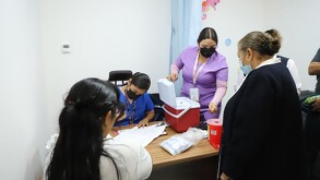 Dos enfermeras con mascarillas coordinan una jornada de vacunación en un consultorio; una revisa documentos y la otra organiza una hielera roja.