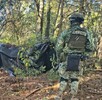 Dos soldados del Ejército Mexicano, de espaldas y con equipo táctico, vigilan un campamento improvisado con lonas negras en una zona boscosa.