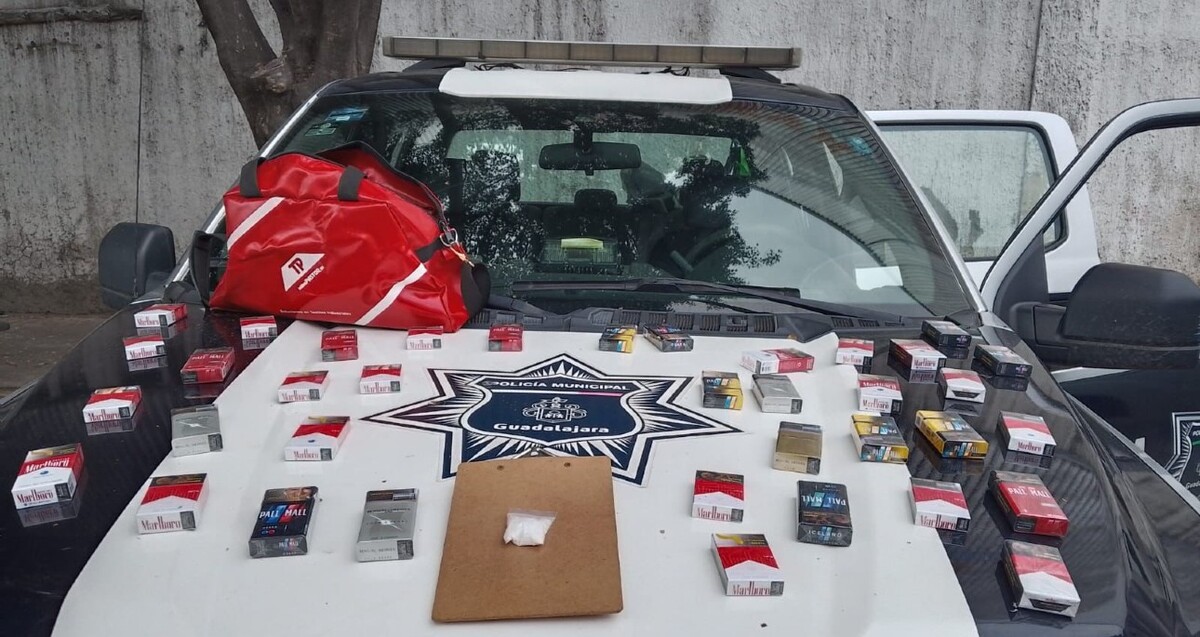 36 cajetillas de cigarro y un sobre con droga sobre el cofre de una patrulla de la Policía de Guadalajara con las puertas abiertas