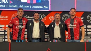 Cuatro hombres posan en una mesa de prensa del Atlas FC; dos jugadores visten la camiseta rojinegra y otros dos directivos usan chamarras del club.