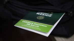Una Cartilla Nacional de Salud de México, de color verde, sobresale del bolsillo de una mochila negra, enfocando los datos de vacunación.