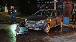 Automóvil color negro con la fascia desprendida luego de un choque en medio de la calle mojada y con personas alrededor