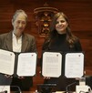 Dos mujeres muestran documentos oficiales firmados frente al escudo de la Universidad de Guadalajara, con una bandera de México al costado.
