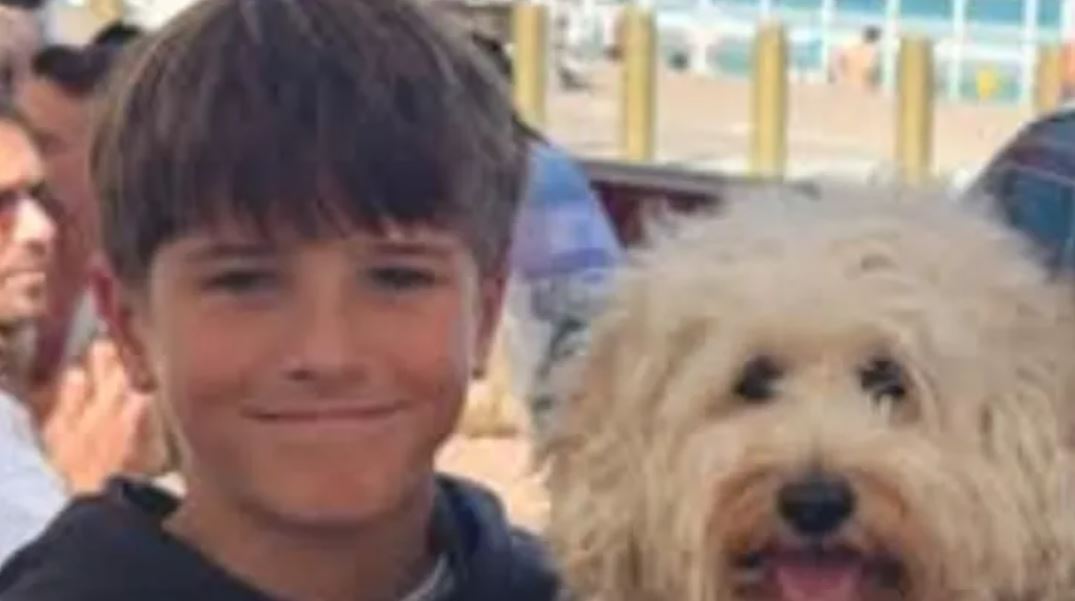 Niño identificado como Nico Antic sonriendo al lado de un perro con personas caminando y una playa de fondo