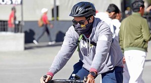 Hombre con la cara cubierta con una pañoleta y lentes circulando en bicicleta con personas de fondo durante un día soleado