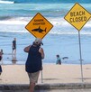 Adultos y niños caminando frente a letreros de alerta por tiburones y el cierre de playas frente al mar