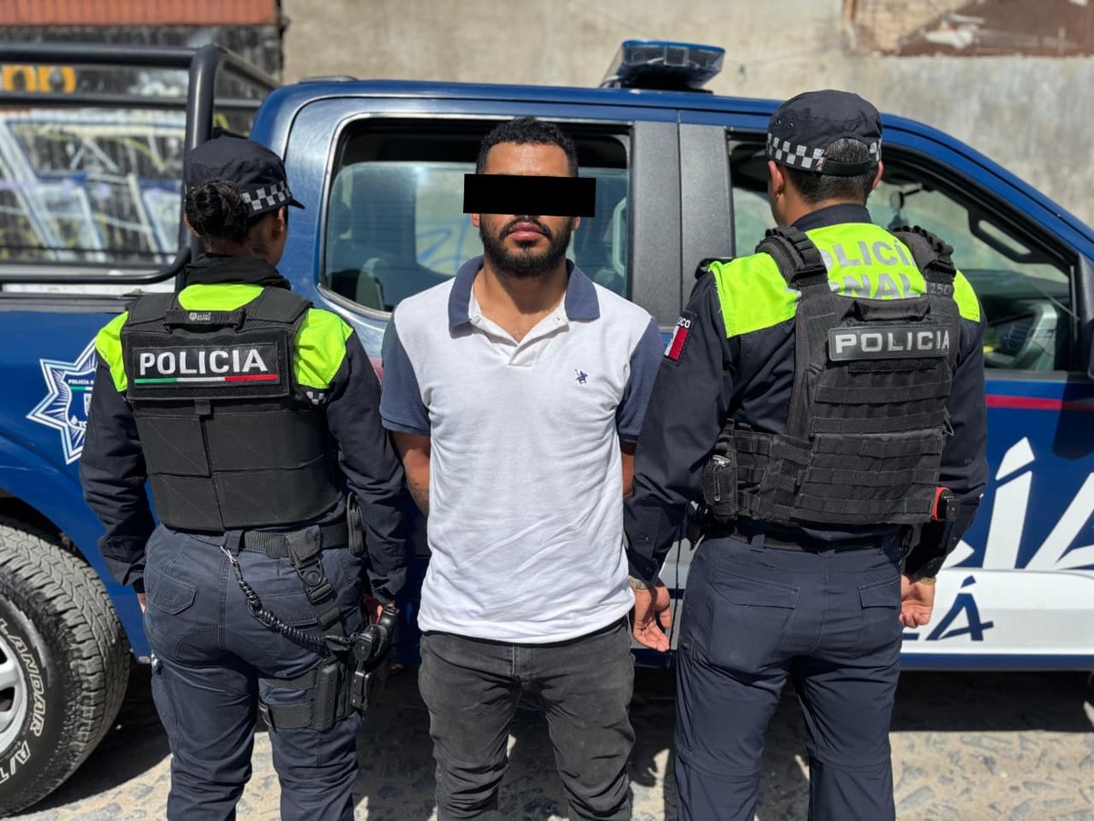 Hombre viendo de frente con los ojos censurados por un recuadro negro en medio de dos policías de espaldas frente a una patrulla de Tonalá