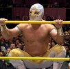 El luchador Místico, con su máscara plateada y dorada, se pone de cuclillas sobre las cuerdas de un ring de lucha libre frente a una multitud.