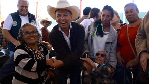 Pablo Lemus, con sombrero de charro, sonríe y estrecha la mano de una mujer adulta mayor durante un evento público rodeado de varios ciudadanos.