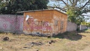 Una pequeña construcción de color naranja con una antena parabólica, cuyas paredes exteriores muestran diversos grafitis con siglas y letras.