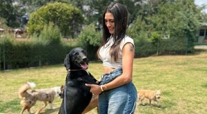 Mujer sosteniendo de las patas a un perro color negro mientas sonríe con otros perros paseando en el fondo en medio de un jardín