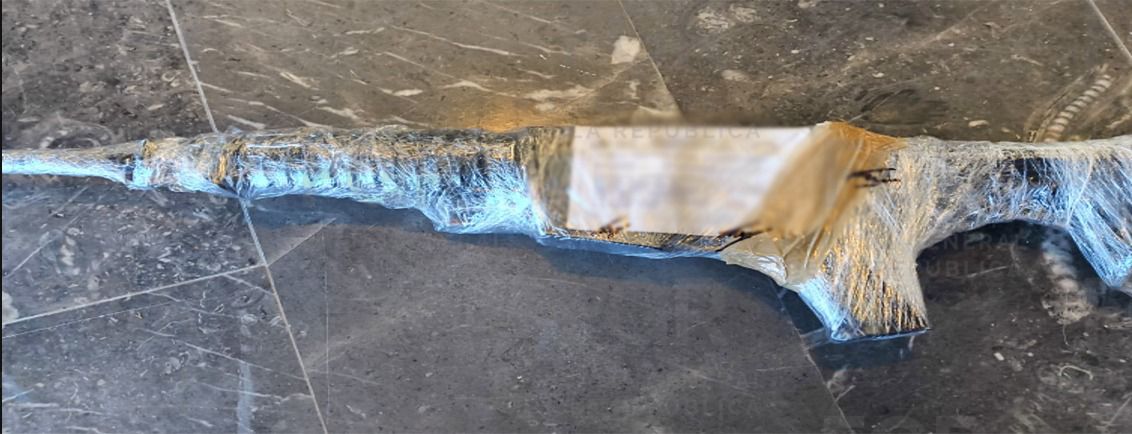 Arma larga tipo fusil envuelta en plástico colocada sobre el piso de loseta negra tras ser asegurada por la FGR