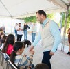 El político Quirino Ordaz Coppel, de camisa azul y chaleco color crema, saluda y choca los cinco con una niña en un evento al aire libre bajo una carpa, rodeado