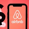 Celular con la aplicación de Airbnb abierta al lado de la sombra de una mano con el signo de pesos delante de un letrero de Airbnb