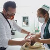 Una enfermera del IMSS y un padre de familia, ambos con mascarillas, atienden a un recién nacido sobre una mesa de auscultación en un consultorio.
