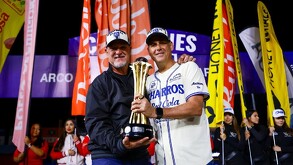 Dos hombres sonrientes con gorras de campeones sostienen juntos un trofeo dorado en un campo de béisbol por la noche, rodeados de banderas de colores.