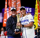 Dos hombres sonrientes con gorras de campeones sostienen juntos un trofeo dorado en un campo de béisbol por la noche, rodeados de banderas de colores.