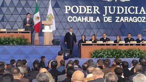 Se observa una persona parada en un escenario, hay una mujer que sostiene una hoja en sus manos abajo del escenario.