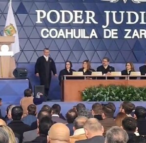 Se observa una persona parada en un escenario, hay una mujer que sostiene una hoja en sus manos abajo del escenario.