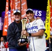 Dos hombres sonrientes con gorras de campeones sostienen juntos un trofeo dorado en un campo de béisbol por la noche, rodeados de banderas de colores.
