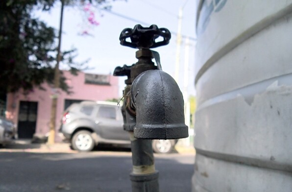 Primer plano de una llave de paso metálica conectada a un gran tinaco blanco en una calle residencial, con autos y casas de fondo desenfocados.