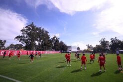 Jugadores de las Chivas de Guadalajara entrenan en una cancha de césped verde bajo un cielo despejado, frente a una barda roja con el nombre del club.