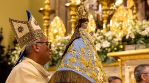 Padre sosteniendo una figura de la Virgen de San Juan de los Lagos dentro de la Basílica con adornos y personas alrededor