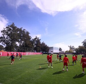 Jugadores de las Chivas de Guadalajara entrenan en una cancha de césped verde bajo un cielo despejado, frente a una barda roja con el nombre del club.