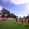Jugadores de las Chivas de Guadalajara entrenan en una cancha de césped verde bajo un cielo despejado, frente a una barda roja con el nombre del club.