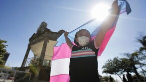 Niña usando un cubrebocas y levantando la bandera trans a contraluz con un kiosko, árboles y personas de fondo