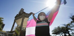 Niña usando un cubrebocas y levantando la bandera trans a contraluz con un kiosko, árboles y personas de fondo