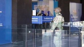 Se observa a un hombre con uniforme de militar y con el rostro cubierto, afuera de un edificio con cartelones azules.