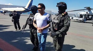 César Alejandro, alias El Bótox siendo sujetado por dos oficiales de la SSPC con un avión y un helicóptero detrás y un camarógrafo a la izquierda