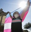 Niña usando un cubrebocas y levantando la bandera trans a contraluz con un kiosko, árboles y personas de fondo