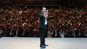 Se observa a un joven parado en un escenario con traje negro tomándose una selfie con un grupo de personas que están atrás.