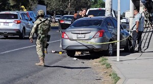 Militar del Ejército al lado de un Nissan Versa color gris acordonado por una cinta policiaca con personas alrededor y autos transitando al lado