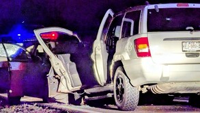 Una camioneta blanca con las puertas abiertas es escoltada por una patrulla con luces rojas y azules encendidas en una carretera durante la noche.