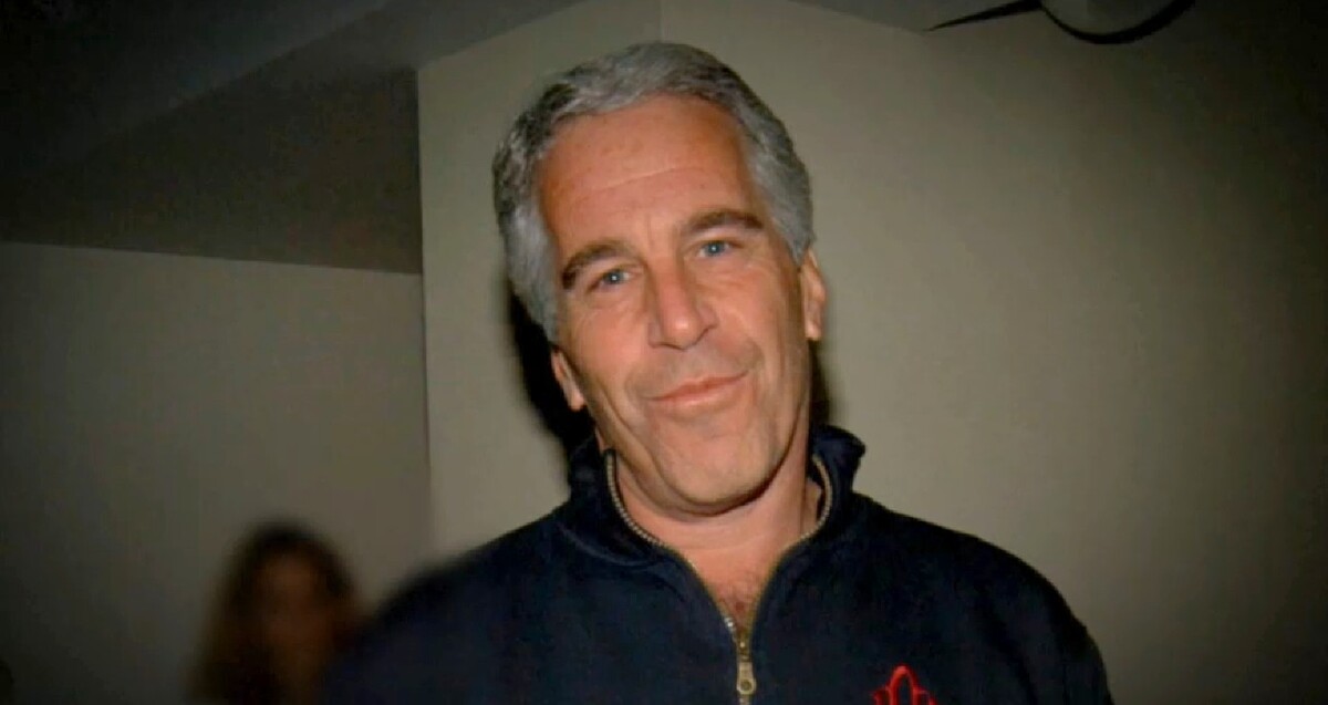 Jeffrey Epstein viendo a cámara mientras sonríe y viste un suéter color azul con una mujer desenfocada en el fondo