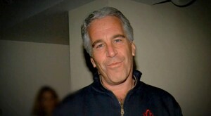 Jeffrey Epstein viendo a cámara mientras sonríe y viste un suéter color azul con una mujer desenfocada en el fondo