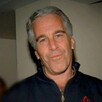 Jeffrey Epstein viendo a cámara mientras sonríe y viste un suéter color azul con una mujer desenfocada en el fondo
