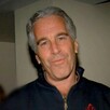Jeffrey Epstein viendo a cámara mientras sonríe y viste un suéter color azul con una mujer desenfocada en el fondo
