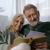 una pareja de adultos mayores, una mujer y u hombre de la tercera edad sentados en una sala mientras leen un libro