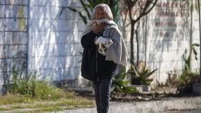 Se ve a una mujer de la terecra edda con cabello canoso y usando bufanda y chamarra, que va caminando sobre la calle.